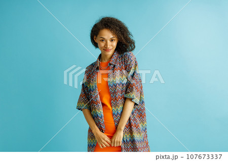 Smiling woman with curly hair in orange dress posing on blue studio background and looks camera 107673337