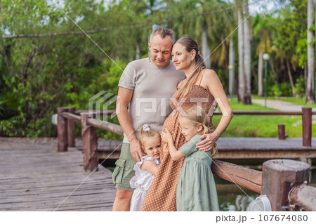A happy, mature couple over 40 with their two daughters, enjoying a leisurely walk in a park, their joy evident as they embrace the journey of pregnancy later in life A happy, mature couple over 40 with their two daughters, enjoying a leisurely walk in a park, their joy evident as they embrace the journey of pregnancy later in life 107674080