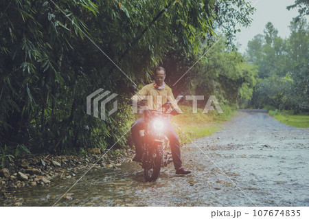 asian man riding small enduro motorcycle crossing flowing creek in forest among rain falling 107674835