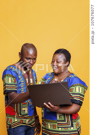 Smiling black man and woman sharing one laptop for work and online pastime activity. Cheerful african american couple wearing ethnic clothes using portable computer in studio 107676077