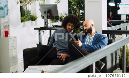 Two employees comparing notes in office, reviewing online company data on laptop and tablet. People planning new presentation for executive board meeting, using official papers. Tripod shot. 107676112