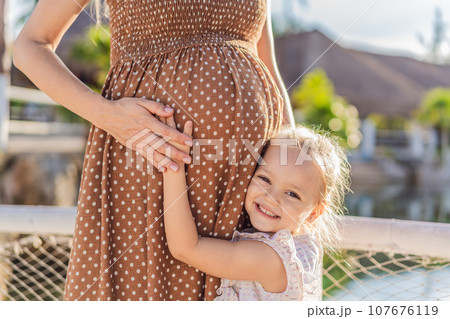A heartwarming moment captured as a daughter lovingly embraces her mother's pregnant belly, sharing in the excitement and anticipation of a new family member A heartwarming moment captured as a daughter lovingly embraces her mother's pregnant belly, sharing in the excitement and anticipation of a new family member 107676119