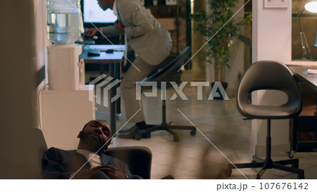 Team leader finishing nightshift, ready to go home, catching tired assistant taking nap on office couch. Manager annoyed by employee slacking off by sleeping in workspace Team leader finishing nightshift, ready to go home, catching tired assistant taking nap on office couch. Manager annoyed by employee slacking off by sleeping in workspace 107676142