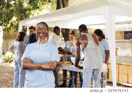 Young black woman with glasses stands outdoors, arms crossed, looking at the camera. Diverse group of volunteers supports a non-profit program dedicated to hunger relief and helping needy individuals. 107676241