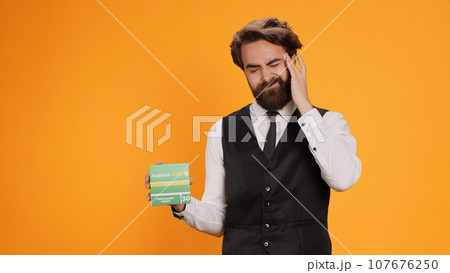 Classy waiter with headache recommends pills on camera, showcasing pack of painkillers to ease pain. Young professional worker feeling unwell and ill, holding medicine treatment box. Classy waiter with headache recommends pills on camera, showcasing pack of painkillers to ease pain. Young professional worker feeling unwell and ill, holding medicine treatment box. 107676250