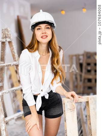 Sensual long-haired woman in hardhat, shirt and shorts posing in room under renovation 107684008