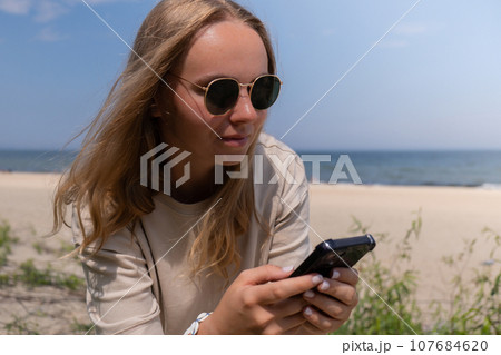 Female hand with shell bracelets holding smartphone. Unrecognizable woman using mobile phone on the beach seacoast. Holiday vacation advertisement. Social media Female hand with shell bracelets holding smartphone. Unrecognizable woman using mobile phone on the beach seacoast. Holiday vacation advertisement. Social media 107684620