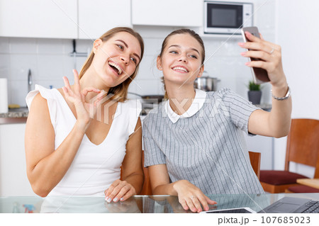Two young girlfriends making selfie by phone and smiling at table 107685023