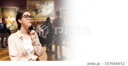 Web banner of art exhibition. Portrait of thoughtful young Caucasian woman wearing glasses and looking at art. In background, people are looking at paintings. Concept of Museum Day Web banner of art exhibition. Portrait of thoughtful young Caucasian woman wearing glasses and looking at art. In background, people are looking at paintings. Concept of Museum Day 107686652