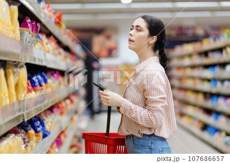 Side view of portrait of pretty caucasian woman holds smartphone and shopping cart. Choice products in supermarket Side view of portrait of pretty caucasian woman holds smartphone and shopping cart. Choice products in supermarket 107686657