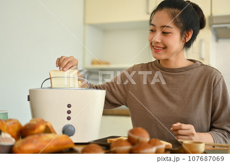Happy young woman toasting bread with toaster in the kitchen. People, food and domestic life concept Happy young woman toasting bread with toaster in the kitchen. People, food and domestic life concept 107687069