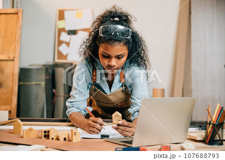 Carpenter america black woman curly hair sketch making notes in work paper while standing at wooden table with laptop computer, young female working learning online at woodshop, Happy Carpenters Day 107688793
