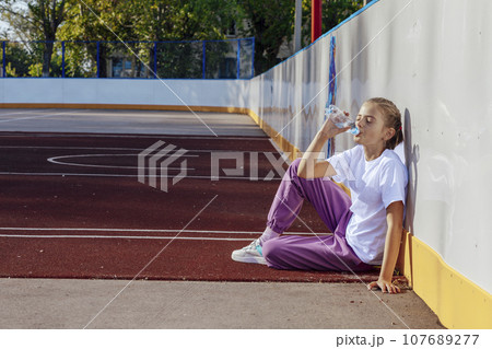 girl drinks water while sitting on a rubber coating on the basketball court. Sport, active lifestyle concept girl drinks water while sitting on a rubber coating on the basketball court. Sport, active lifestyle concept 107689277