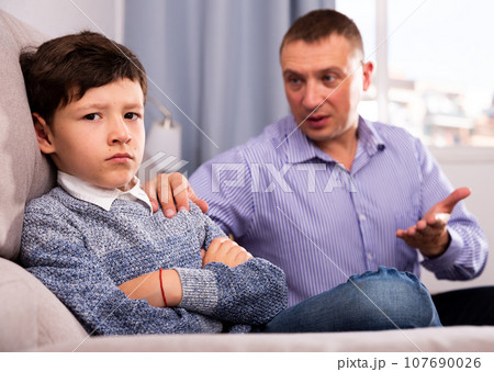 Young father consoling preteen son sitting at sofa after quarrel Young father consoling preteen son sitting at sofa after quarrel 107690026