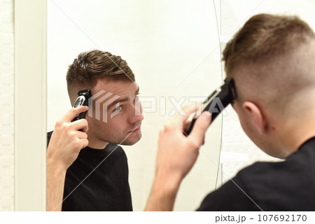Young man trimming hair and looking in the mirror in a bathroom Young man trimming hair and looking in the mirror in a bathroom 107692170