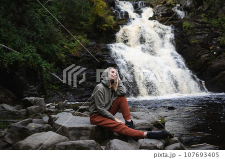 a young beautiful woman tourist sitting against the backdrop of a waterfall surrounded by forest a young beautiful woman tourist sitting against the backdrop of a waterfall surrounded by forest 107693405