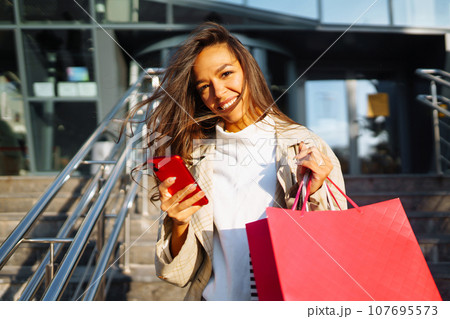 Young woman standing along road holding shopping bags, using mobile phone. Discounts, sale concept. Young woman standing along road holding shopping bags, using mobile phone. Discounts, sale concept. 107695573