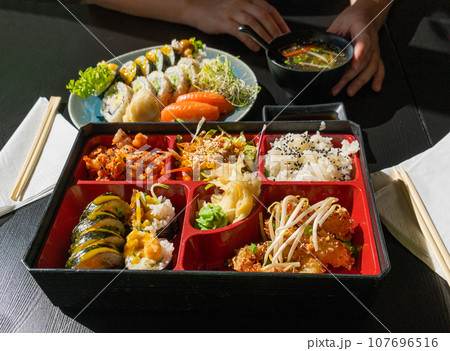 Japanese Lunch in Bento Box with Deep-Fried Squid Rings, Sushi Rolls, Salad, Rice, Pickled Ginger 107696516