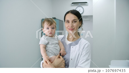 Pediatrician stands in middle of hospital room and looks at camera Pediatrician stands in middle of hospital room and looks at camera 107697435