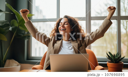 Cute, happy woman with closed eyes sitting at a home office desk with a laptop, raising her arms in a victory pose Cute, happy woman with closed eyes sitting at a home office desk with a laptop, raising her arms in a victory pose 107698895