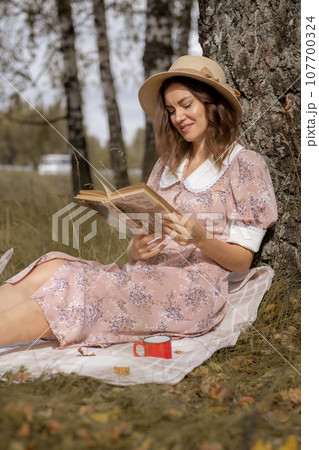A young beautiful woman in a dress and a round hat reads a book outdoors in the forest and drinks tea. Romantic and vintage photo of a beautiful girl. Reading and relaxation A young beautiful woman in a dress and a round hat reads a book outdoors in the forest and drinks tea. Romantic and vintage photo of a beautiful girl. Reading and relaxation 107700324