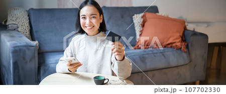 Smiling cute asian woman using credit card and smartphone, paying bills online, holding mobile phone, looking at camera 107702330
