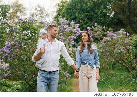 Mother, father and little son are walking in a spring blooming park. A happy mom and dad with a small child are having fun near the lilac. The concept of a happy family. Mother, father and little son are walking in a spring blooming park. A happy mom and dad with a small child are having fun near the lilac. The concept of a happy family. 107705790