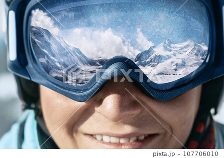 Close up of the ski goggles of a man with the reflection of snowed mountains.  A mountain range reflected in the ski mask.  Man  on the background blue sky. Wearing ski glasses. Winter Sports. 107706010
