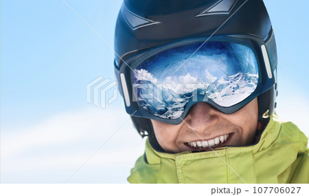 Close up of the ski goggles of a man with the reflection of snowed mountains. A mountain range reflected in the ski mask. Man on the background blue sky. Wearing ski glasses. Winter Sports. Close up of the ski goggles of a man with the reflection of snowed mountains. A mountain range reflected in the ski mask. Man on the background blue sky. Wearing ski glasses. Winter Sports. 107706027