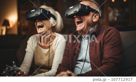 Senior mature couple wearing virtual reality headsets sits on the sofa having fun together. Happy retired people using modern VR goggle glasses. New trends and technology concept for all ages. 107712917