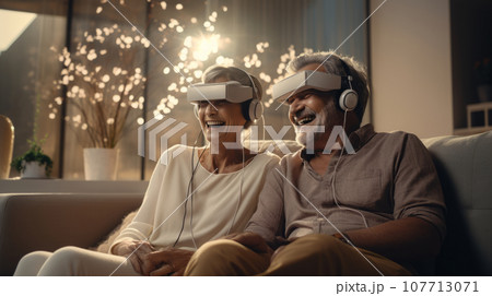 Senior mature couple wearing virtual reality headsets sits on the sofa having fun together. Happy retired people using modern VR goggle glasses. New trends and technology concept for all ages. 107713071