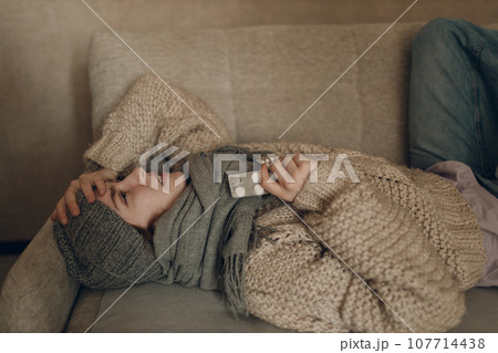 Young woman wearing scarf holding pills in hands has flu ill sick disease cold at home indoor on sofa 107714438