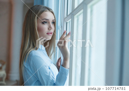 Beautiful woman opening blue curtains looking out window at home Beautiful woman opening blue curtains looking out window at home 107714781