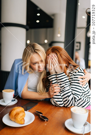 Vertical portrait of two best girlfriends sitting at indoors cafe and talking. Blonde female stroking hand, hugging, trying calm unhappy redhead friend, who cover face with hands and tearing. 107719329