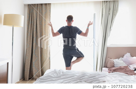 Man stands in lotus position by window Man stands in lotus position by window 107720238