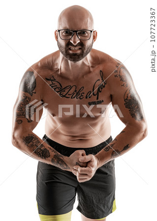 Athletic bald, tattooed man in black shorts is posing isolated on white background. Close-up portrait. Athletic bald, tattooed man in black shorts is posing isolated on white background. Close-up portrait. 107723367