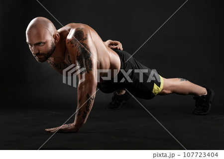 Athletic bald, tattooed man in black shorts and sneakers is posing against a black background. Close-up portrait. Athletic bald, tattooed man in black shorts and sneakers is posing against a black background. Close-up portrait. 107723404