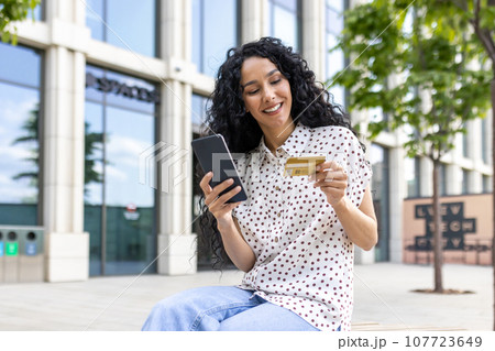 Young beautiful woman walking in the city, satisfied Latin American woman with curly hair holds phone and bank credit card in hands, makes online purchases happily books services and chooses online. Young beautiful woman walking in the city, satisfied Latin American woman with curly hair holds phone and bank credit card in hands, makes online purchases happily books services and chooses online. 107723649