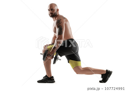 Athletic bald, tattooed man in black shorts and sneakers is posing with a dumbbell isolated on white background. Full length portrait. 107724991