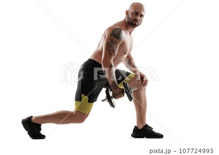 Athletic bald, tattooed man in black shorts and sneakers is posing with a dumbbell isolated on white background. Full length portrait. Athletic bald, tattooed man in black shorts and sneakers is posing with a dumbbell isolated on white background. Full length portrait. 107724993