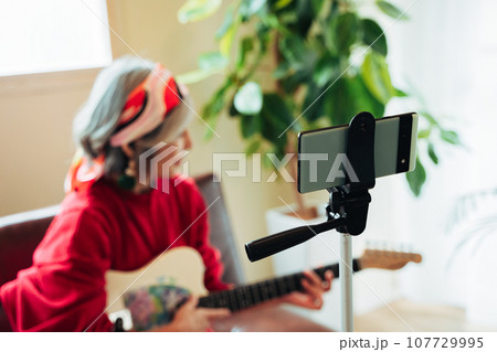 ギター演奏をしながらライブ配信するシニア女性 107729995