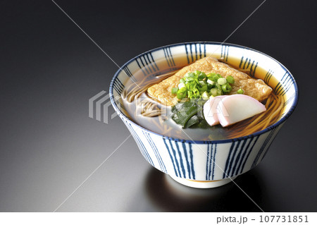 油揚げ、かまぼこ、ワカメ、葱が乗ったきつね蕎麦を黒背景で撮影 107731851