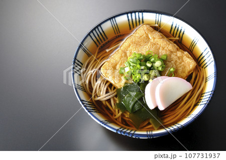 油揚げ、かまぼこ、ワカメ、葱が乗ったきつね蕎麦を黒背景で俯瞰撮影 107731937