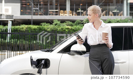Progressive businesswoman leaning on electric car and charging station. 107734590