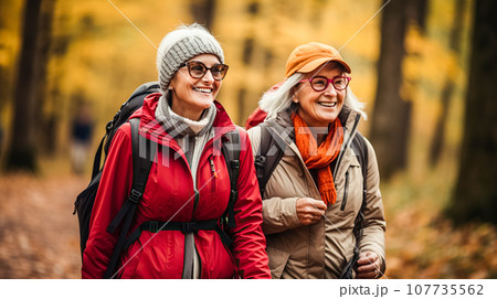 Positive, cheerful elderly women walk with backpacks through autumn forest park. Happy mature ladies travel in group 107735562