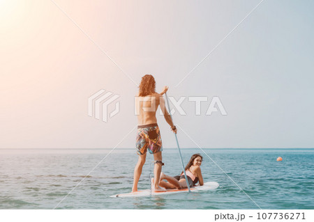 Sea woman and man on sup. Silhouette of happy young woman and man, surfing on SUP board, confident paddling through water surface. Idyllic sunset. Active lifestyle at sea or river. 107736271