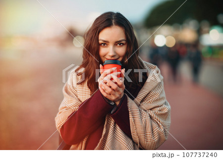 Young smiling woman dressed in autumn coat and plaid drinking hot coffee drink from cardboard cup while strolling outside. 107737240