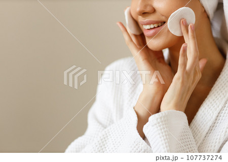 Young woman in white robe and towel on her head wipes the skin of her face with cotton pads. Cleansing and healthy body care after shower. Close-up of pretty girl on isolated background, copy space. Young woman in white robe and towel on her head wipes the skin of her face with cotton pads. Cleansing and healthy body care after shower. Close-up of pretty girl on isolated background, copy space. 107737274