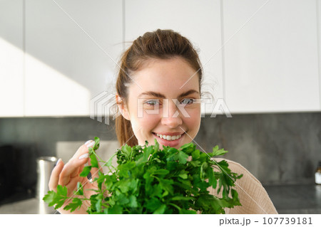 Portrait of beautiful smiling girl with bouquet of parsley, standing in the kitchen and cooking, adding herbs to healthy fresh salad or meal, preparing food 107739181