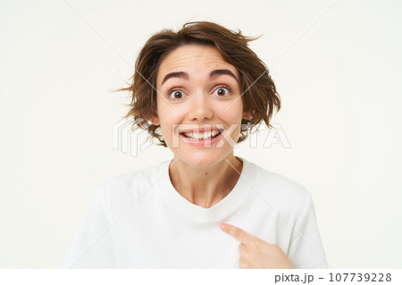 Portrait of girl with surprised face, points at herself amazed, stands isolated over white studio background 107739228